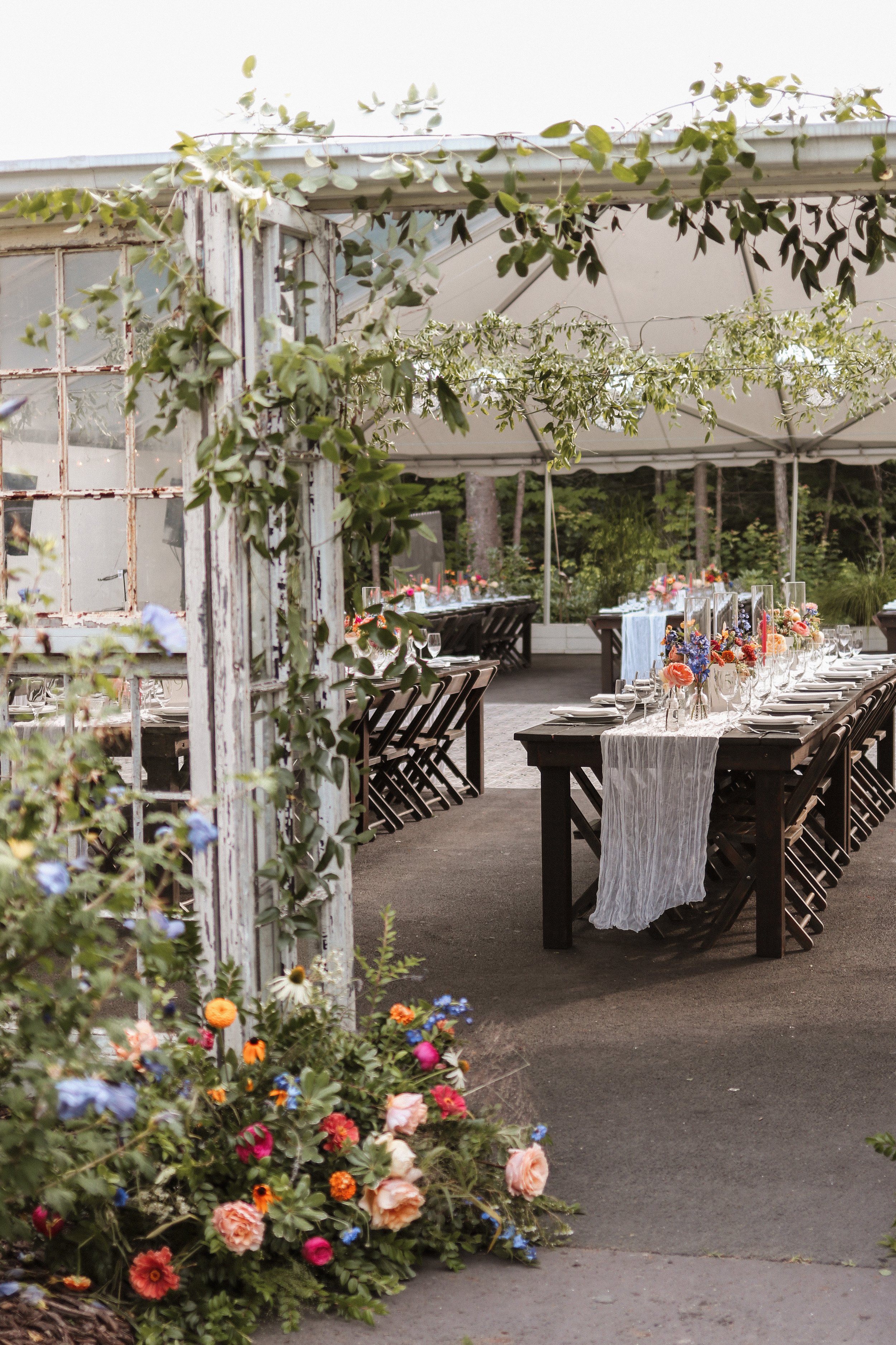 Outdoor wedding reception area with decorated tables, floral arrangements, and white canopy, surrounded by greenery and flowers.